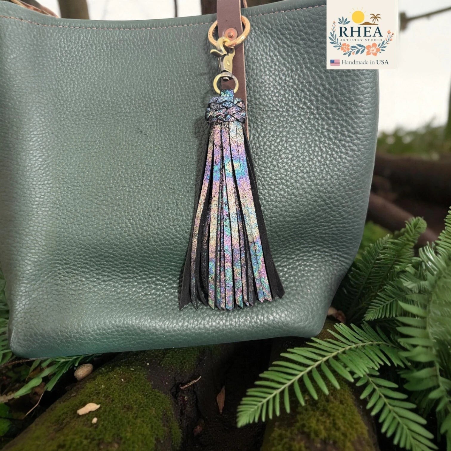 Rhea Artistry Studio handmade double braid leather tassel keychain with iridescent sparkle and antique brass lobster clasp attached to a green pebbled leather bag.