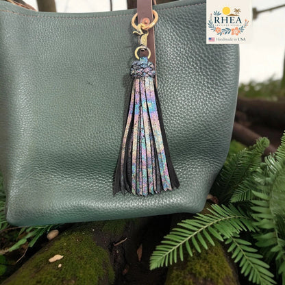 Rhea Artistry Studio handmade double braid leather tassel keychain with iridescent sparkle and antique brass lobster clasp attached to a green pebbled leather bag.
