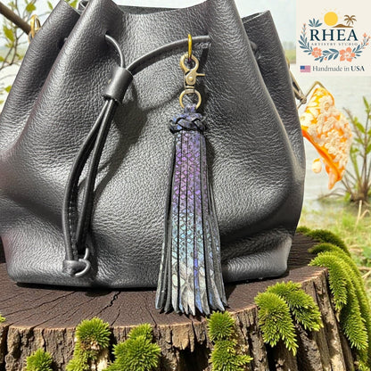 Rhea Artistry Studio handmade large leather tassel keychain in iridescent purple and blue with a lobster clasp, attached to a black leather bag.