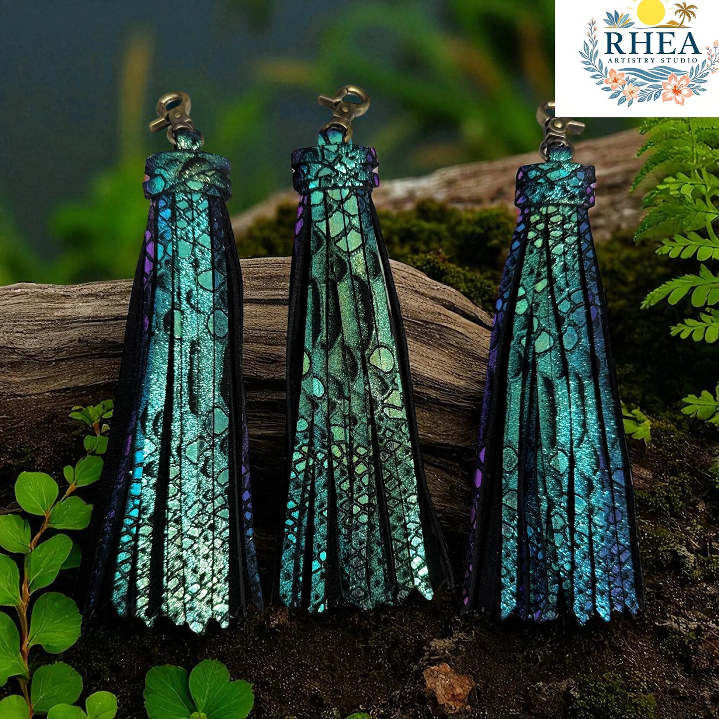 Rhea Artistry Studio handmade teal and purple iridescent leather tassel keychain with lobster clasp, set against a natural wood and foliage background.