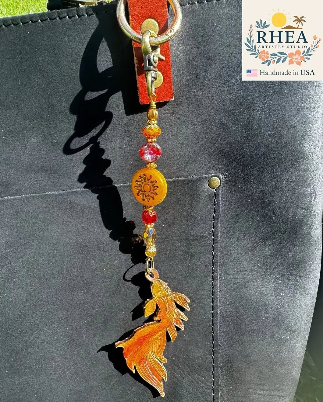 Rhea Artistry Studio orange koi fish leather charm with brass hardware and Czech glass beads on a black bag.