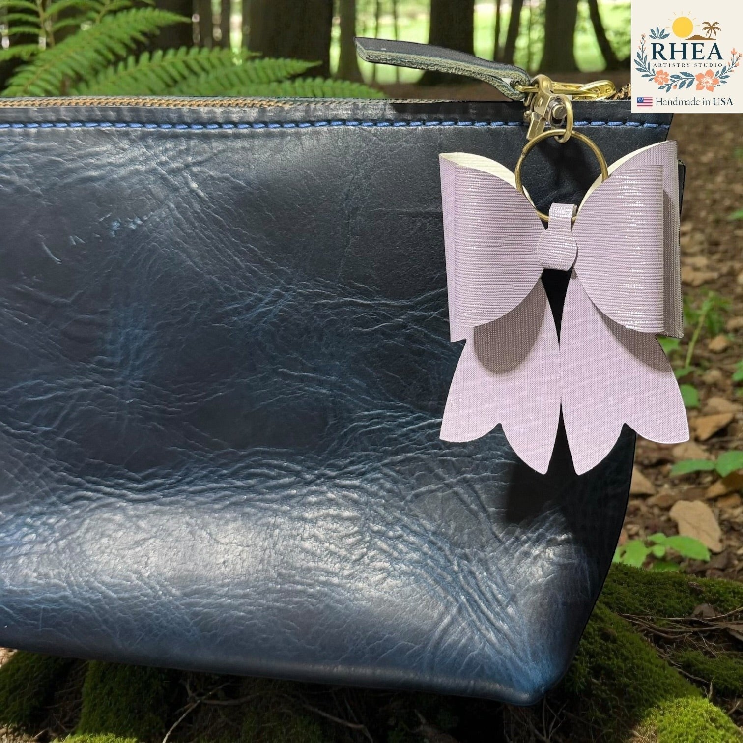 Rhea Artistry Studio lavender textured double-layer French bow bag charm with brass hardware, clipped to a dark blue leather pouch.