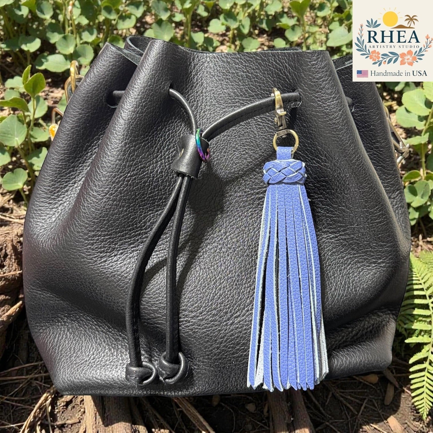 Rhea Artistry Studio lavender leather tassel bag charm with braided detail and brass clasp, attached to a black leather drawstring bag.