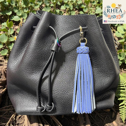 Rhea Artistry Studio lavender leather tassel bag charm with braided detail and brass clasp, attached to a black leather drawstring bag.