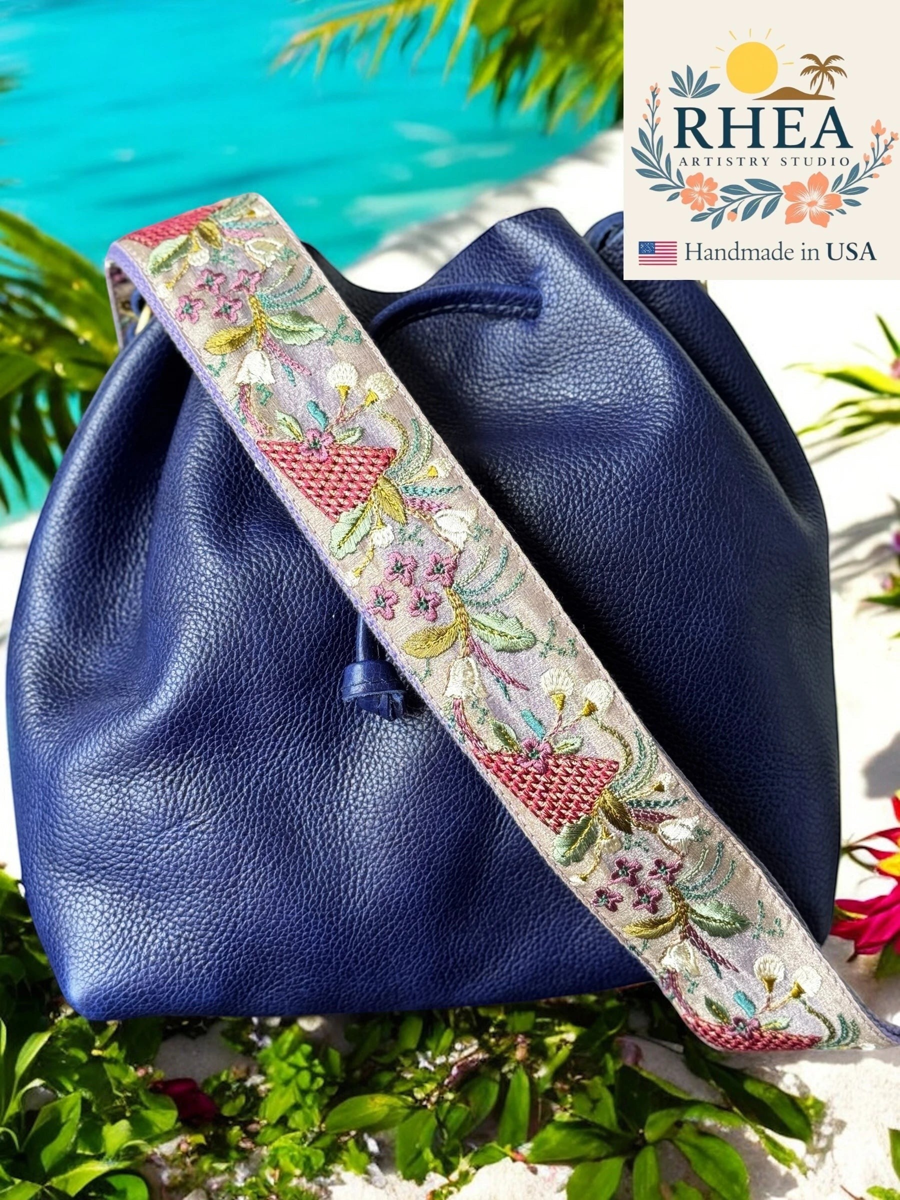Rhea Artistry Studio lavender and purple embroidered crossbody strap with floral and basket motifs on a navy blue leather bag.