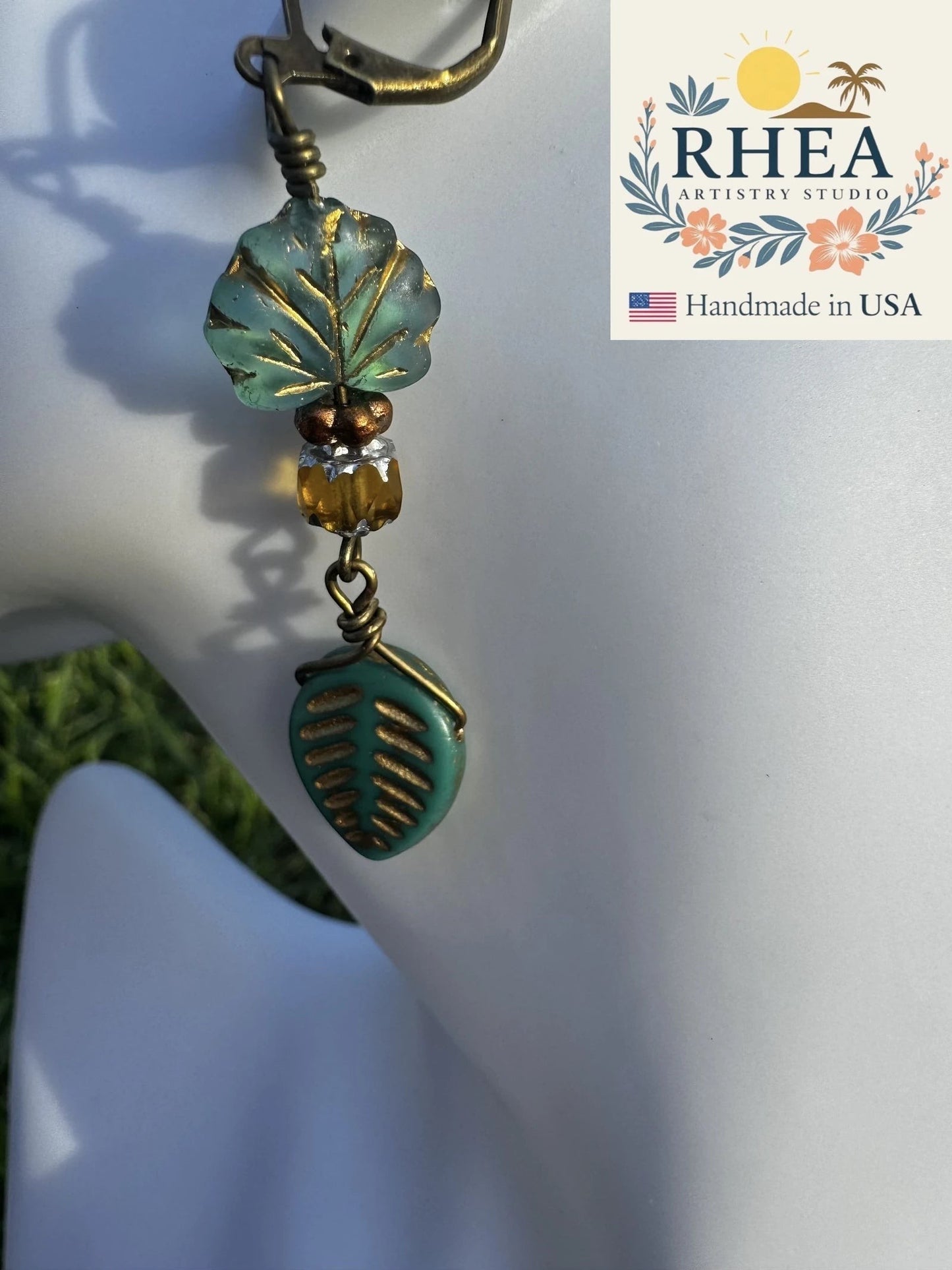 Rhea Artistry Studio handmade earrings with blue-green glass leaf charms and brass hardware, approx. 2 inches long.