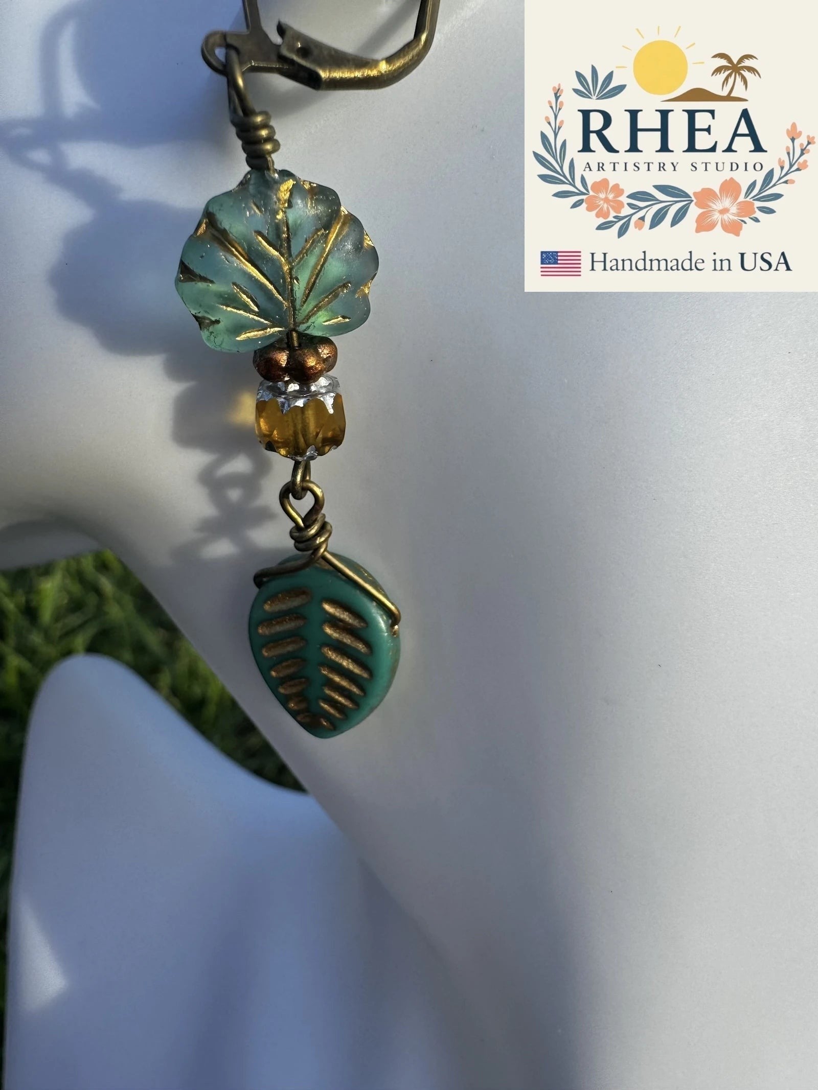 Rhea Artistry Studio handmade earrings with blue-green glass leaf charms and brass hardware, approx. 2 inches long.