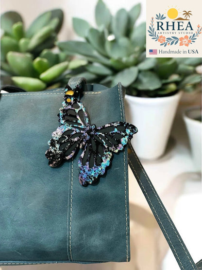 Rhea Artistry Studio black leather butterfly charm with iridescent and metallic finish, attached to a teal purse.