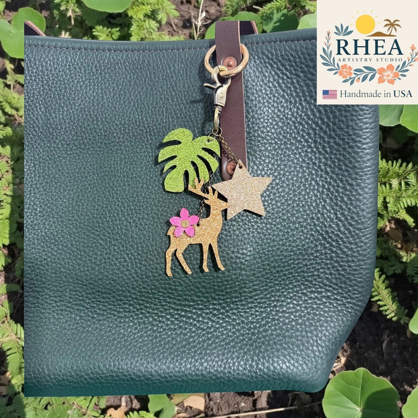 Rhea Artistry Studio handmade leather charm with deer, monstera leaf, and glitter star on a dark green pebbled leather tote bag.
