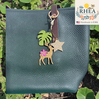 Rhea Artistry Studio handmade leather charm with deer, monstera leaf, and glitter star on a dark green pebbled leather tote bag.