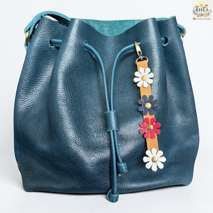 Rhea Artistry Studio handmade leather daisy bag charm with white, navy, red, and white flowers on a tan strap.