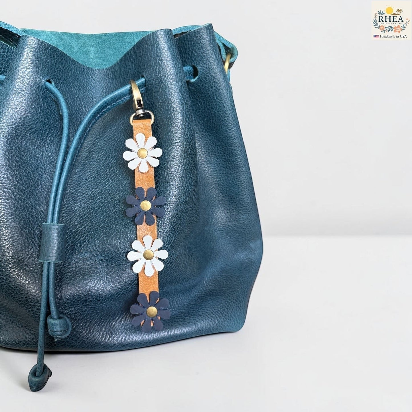 Rhea Artistry Studio handmade leather daisy bag charm with white and blue flowers and brass rivets.
