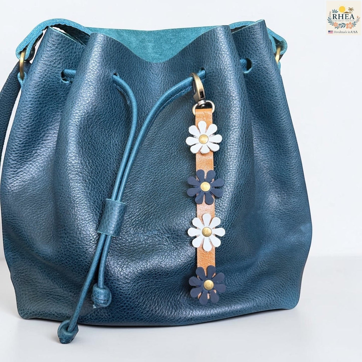 Rhea Artistry Studio handmade leather daisy bag charm with white and blue flowers and brass rivets.