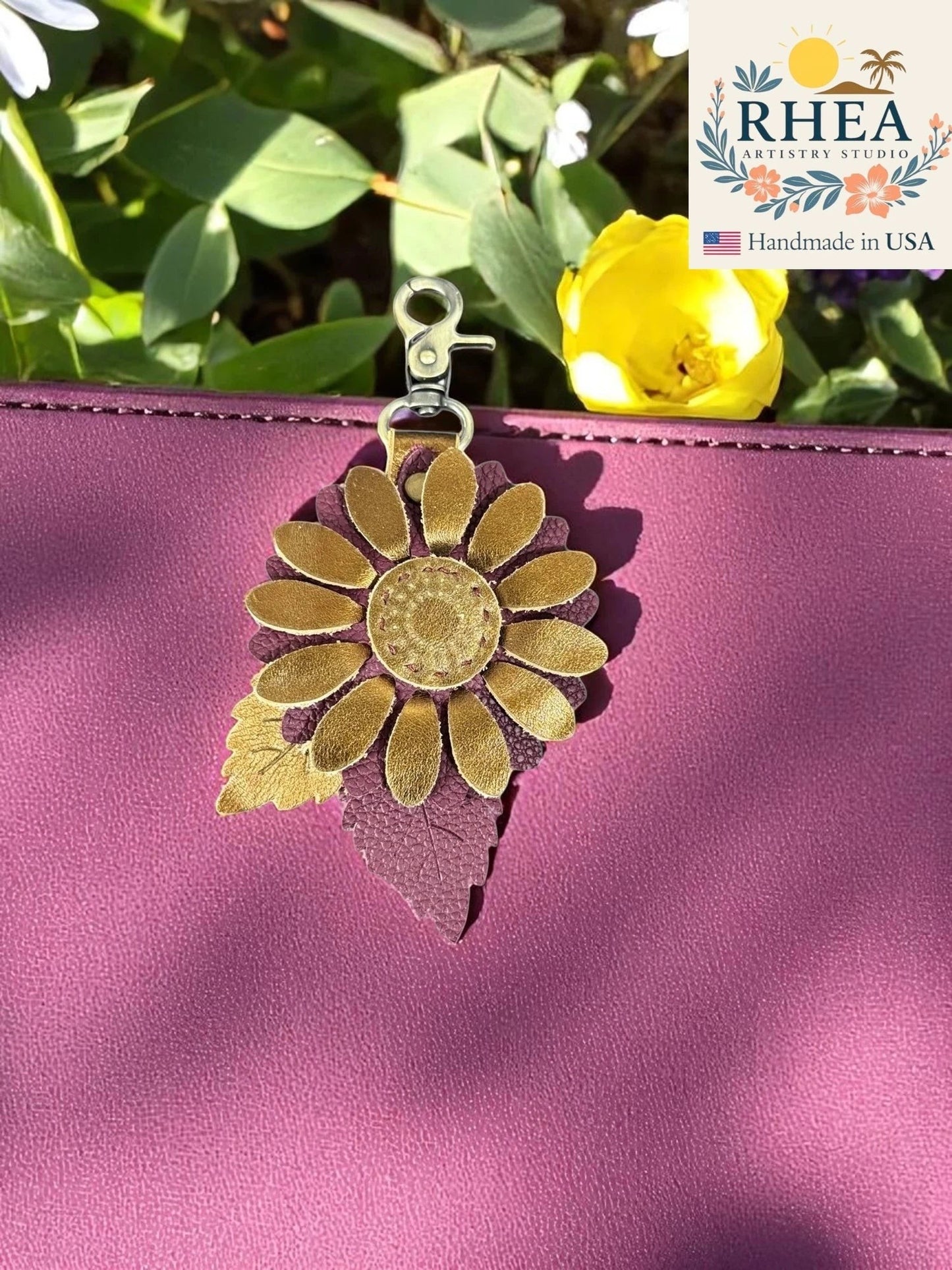Rhea Artistry Studio daisy drop leather charm with gold petals and purple leaf detail on a plum bag.