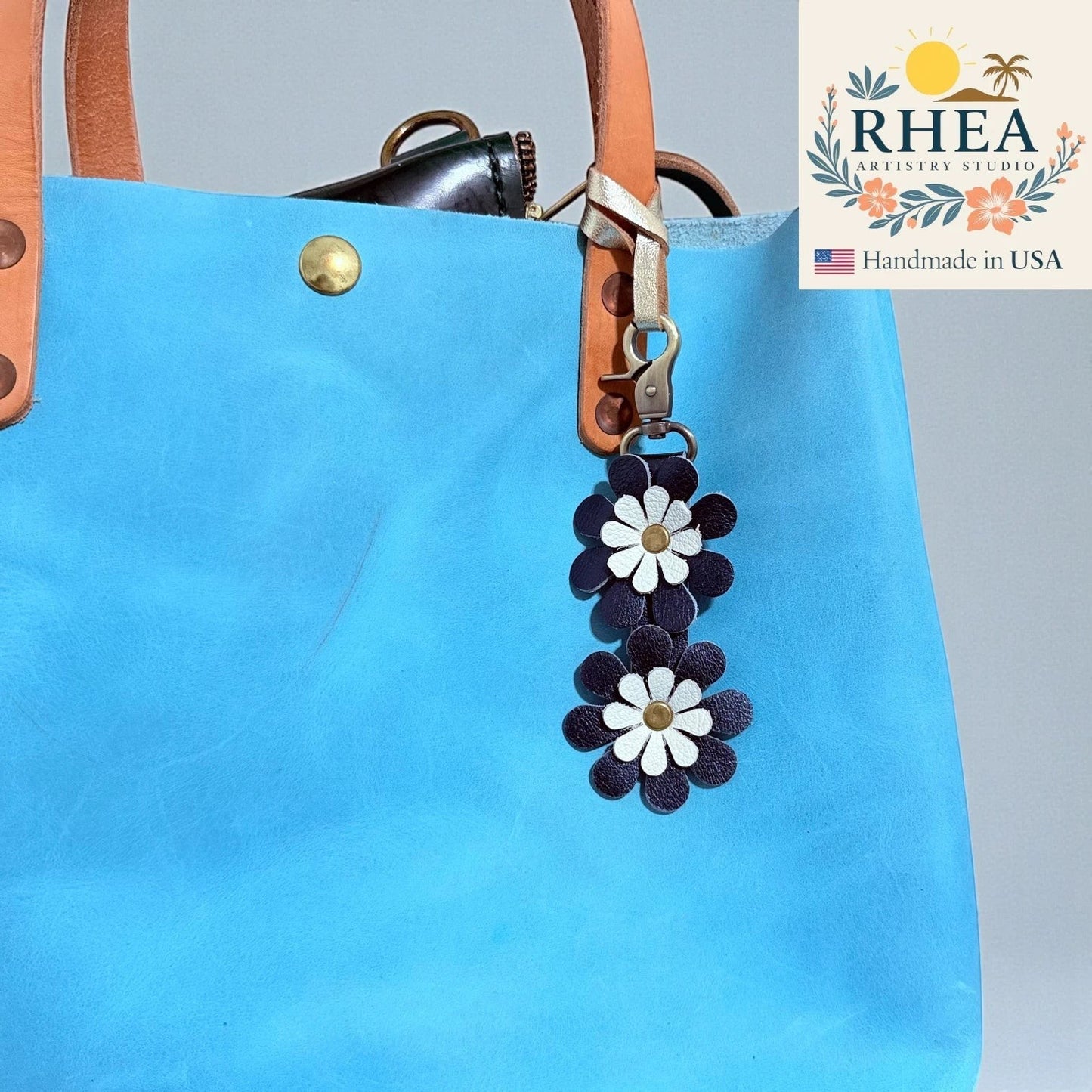 Rhea Artistry Studio handmade double-layer leather daisy charm in navy and white with brass hardware, attached to a bright blue tote bag.