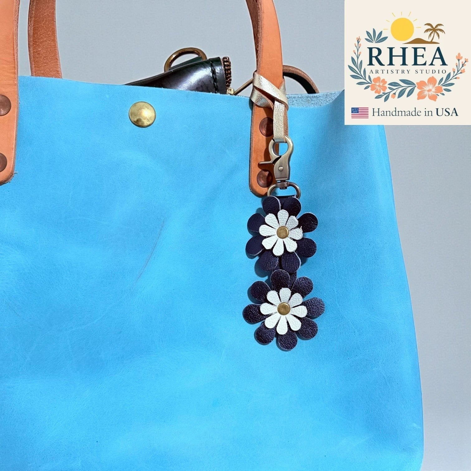 Rhea Artistry Studio handmade double-layer leather daisy charm in navy and white with brass hardware, attached to a bright blue tote bag.