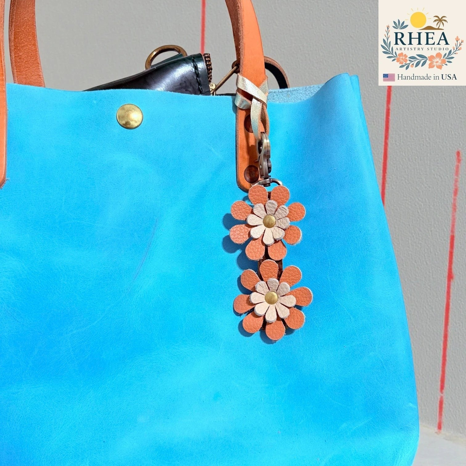 Rhea Artistry Studio handmade double-layer leather daisy charm in orange and cream, attached to a bright blue tote bag.