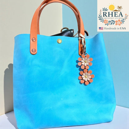 Rhea Artistry Studio handmade double-layer leather daisy purse charm in orange and cream, clipped to a bright blue tote bag.
