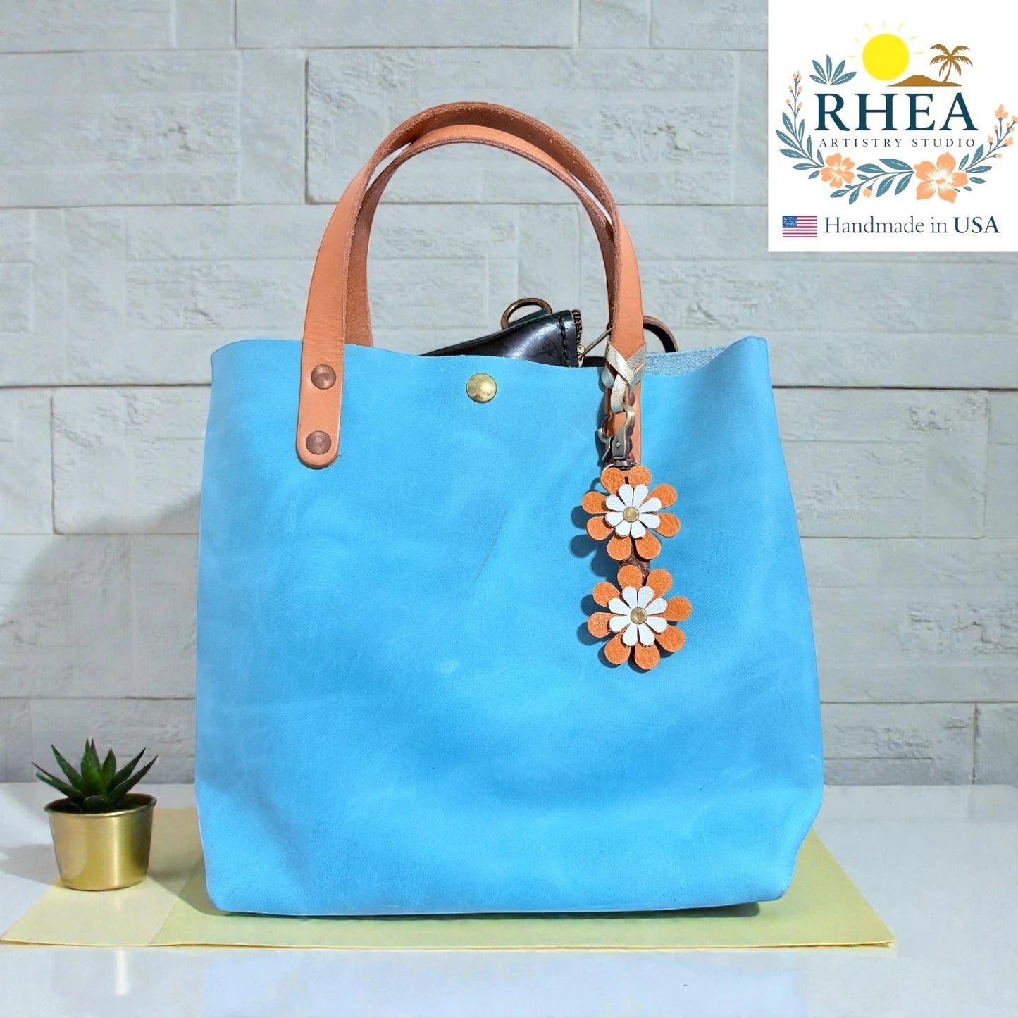 Rhea Artistry Studio double-layer leather daisy purse charm in orange and white, attached to a blue tote bag.