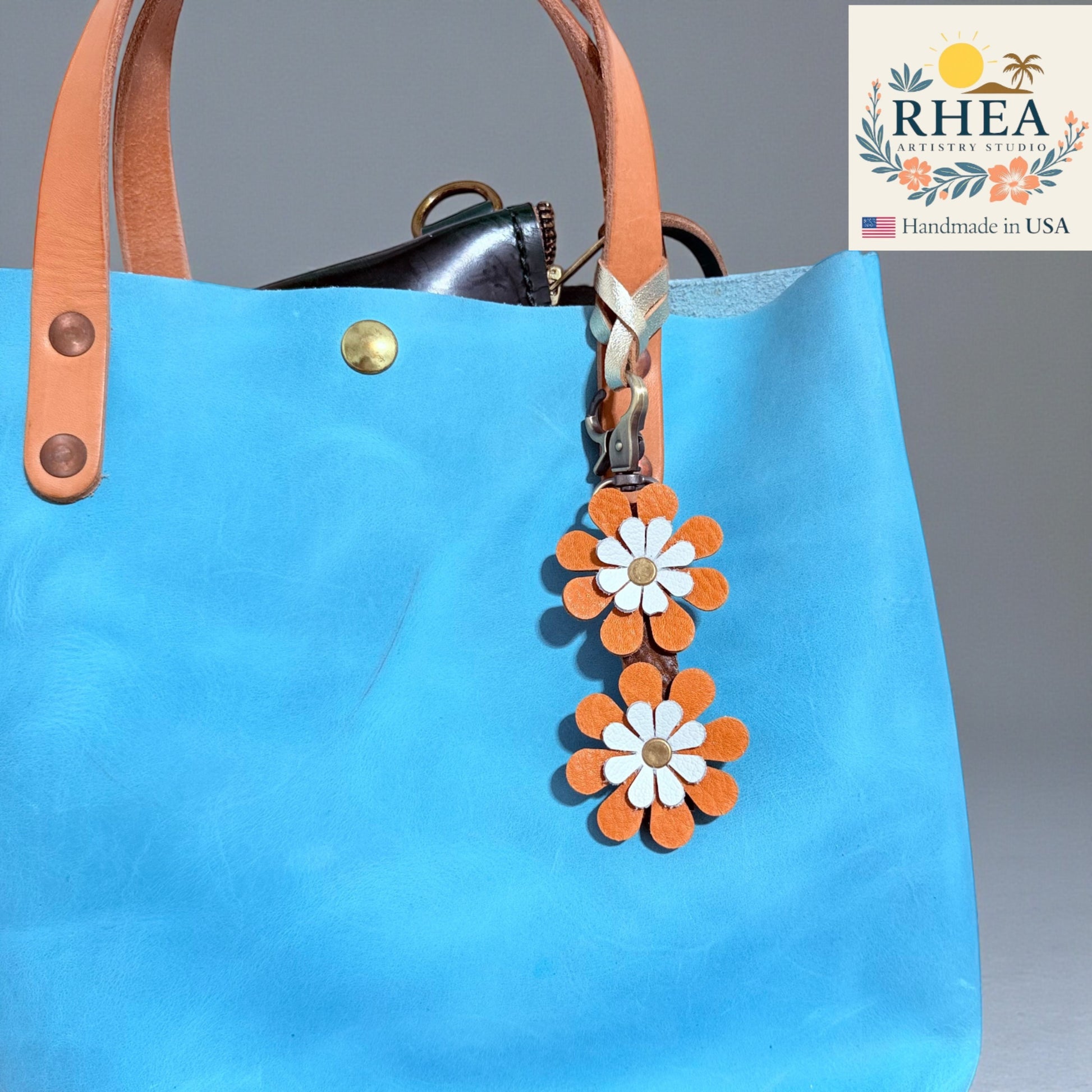 Rhea Artistry Studio handmade double-layer leather daisy charm with orange petals and white centers, attached to a blue bag.