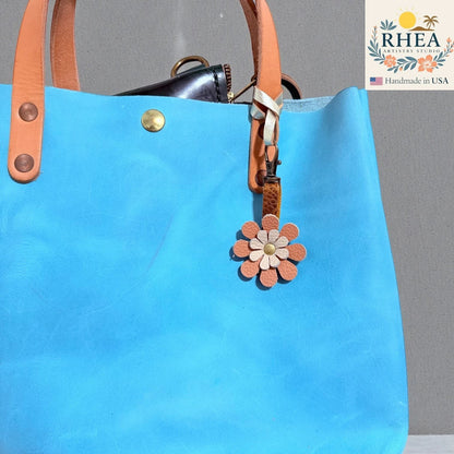 Rhea Artistry Studio handmade double-layer leather daisy charm in peach and cream with brass hardware, attached to a blue tote bag.