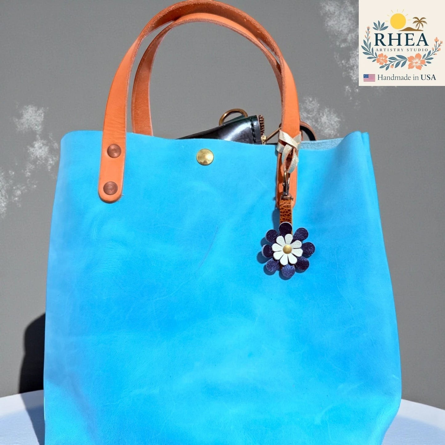 Rhea Artistry Studio handmade double-layer leather daisy charm with dark purple petals and white center, attached to a bright blue tote bag.