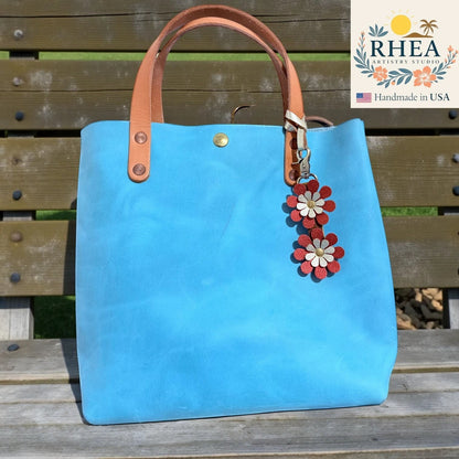 Rhea Artistry Studio handmade double-layer leather daisy purse charm in red and gold, attached to a blue tote bag.