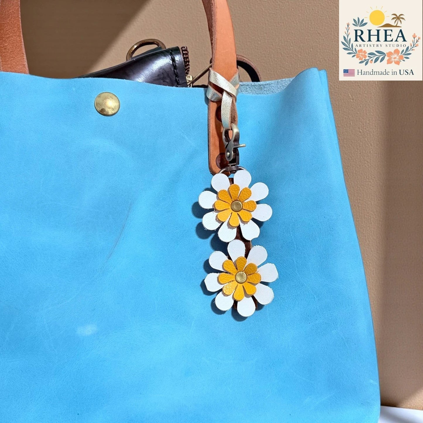 Rhea Artistry Studio handmade double-layer leather daisy charm with white petals and yellow centers, attached to a blue tote bag.
