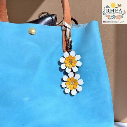 Rhea Artistry Studio handmade double-layer leather daisy charm with white petals and yellow centers, attached to a blue tote bag.