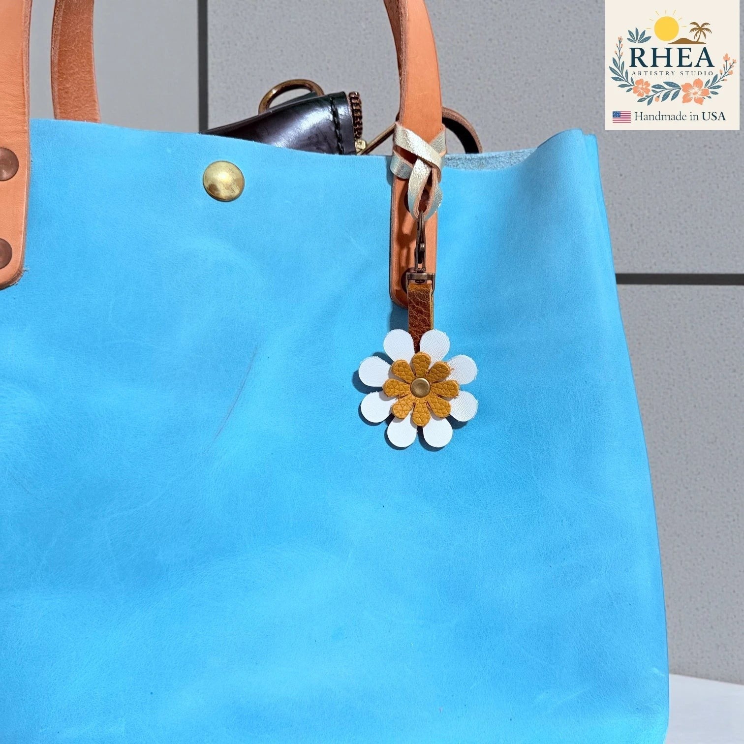 Rhea Artistry Studio handmade double-layer leather daisy charm with white petals and a mustard yellow center, clipped to a blue tote bag.