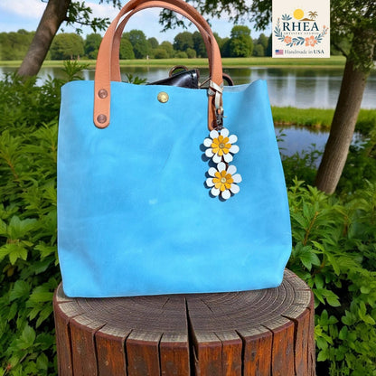 Rhea Artistry Studio handmade double-layer leather daisy charms in white and yellow, attached to a bright blue tote bag.