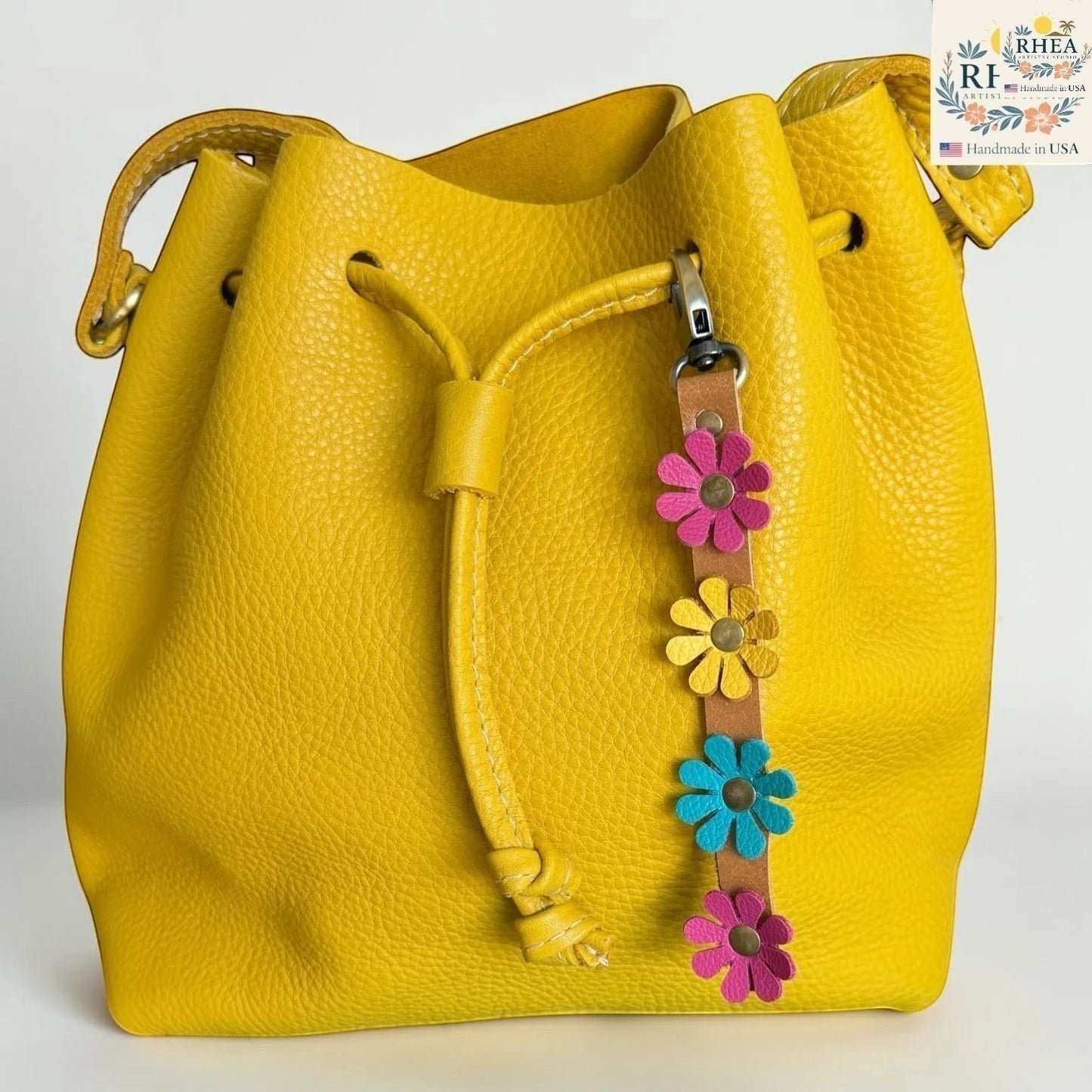 Rhea Artistry Studio handmade leather daisy wristlet keychain with pink, yellow, and blue flowers attached to a tan leather strap, clipped to a yellow leather bucket bag.