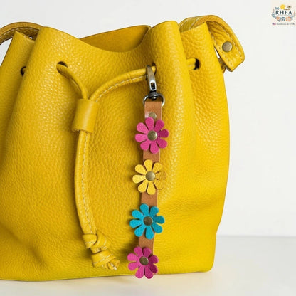 Rhea Artistry Studio handmade leather daisy wristlet keychain with pink, yellow, and blue flowers attached to a tan leather strap with brass rivets, clipped to a yellow leather bucket bag.