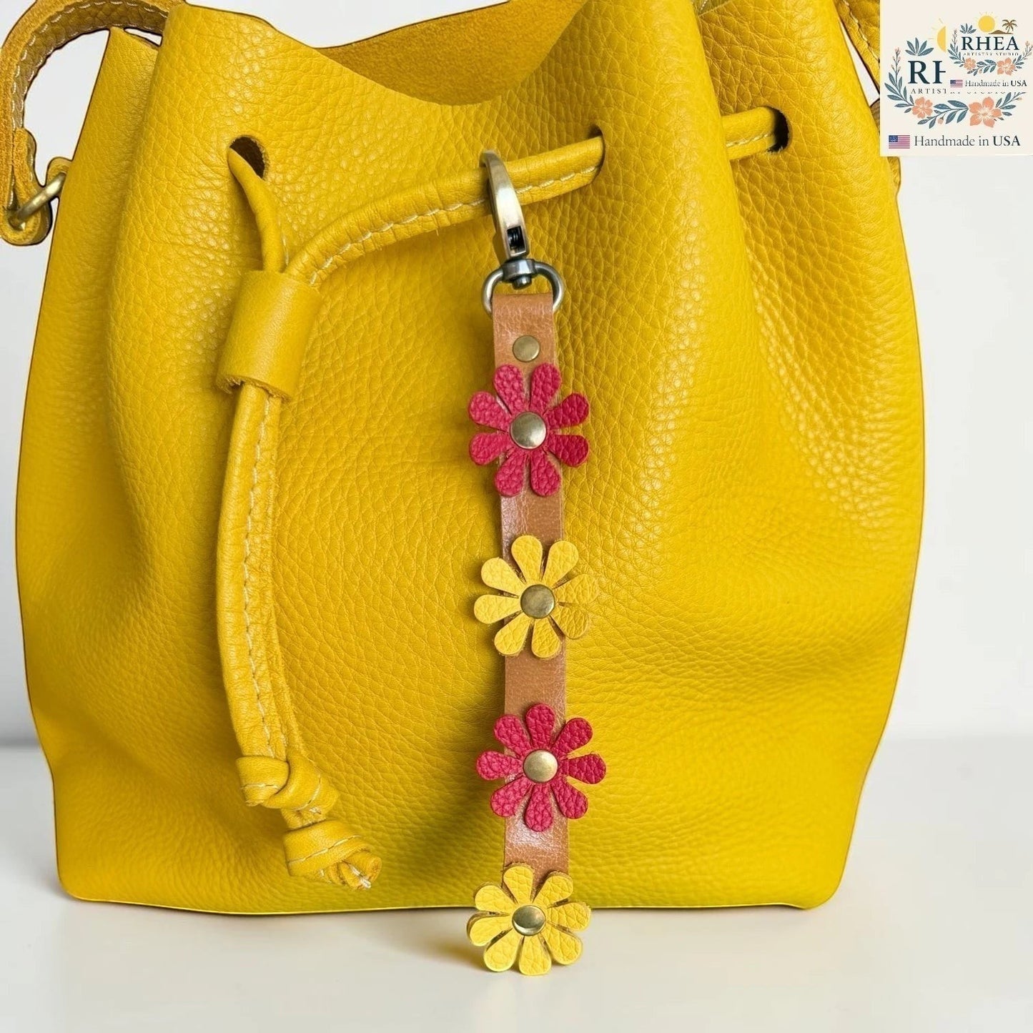 Rhea Artistry Studio handmade leather wristlet keychain with pink and yellow daisy charms and brass rivets, attached to a yellow leather bucket bag.