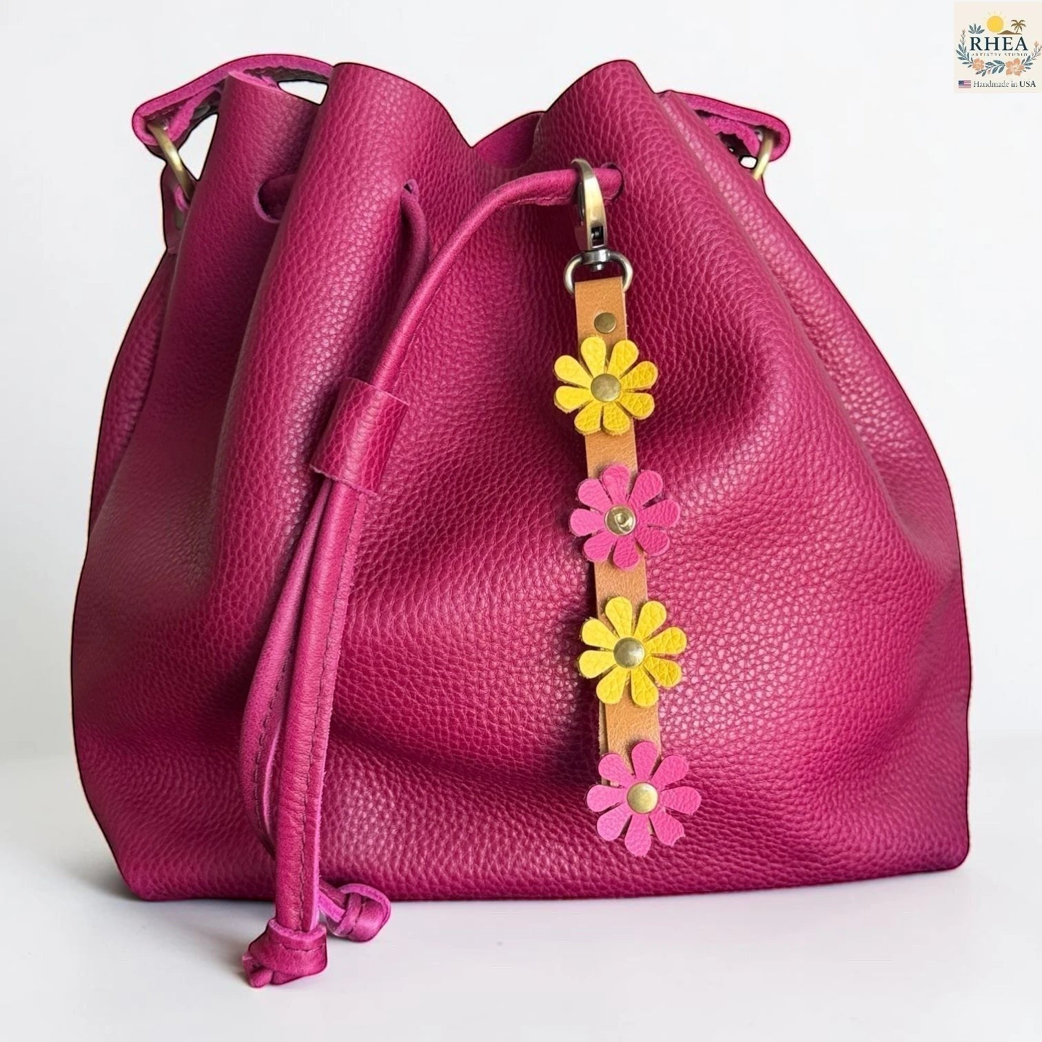 Rhea Artistry Studio handmade leather daisy wristlet keychain with pink and yellow flowers attached to a tan leather strap with brass rivets, clipped to a magenta bucket bag.