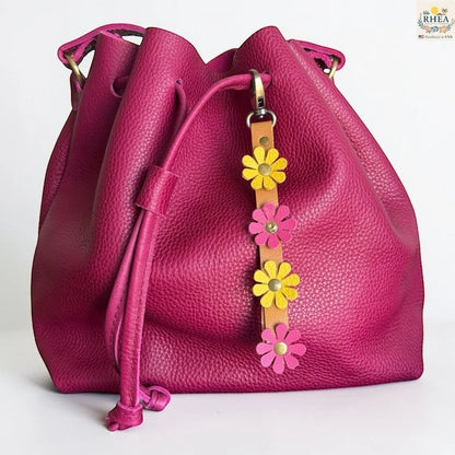 Rhea Artistry Studio handmade leather daisy wristlet keychain with pink and yellow flowers attached to a tan leather strap with brass rivets, clipped to a magenta bucket bag.