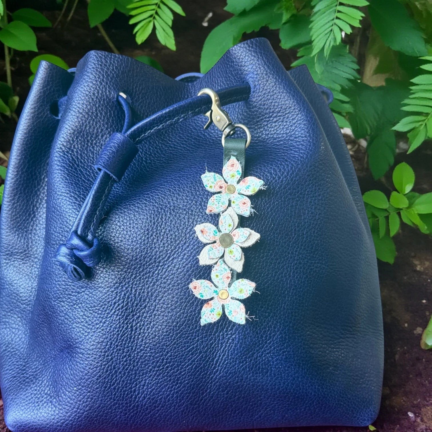 Rhea Artistry Studio navy blue leather bucket bag with a floral charm featuring three fabric flowers with brushed brass centers and a brushed brass clasp.