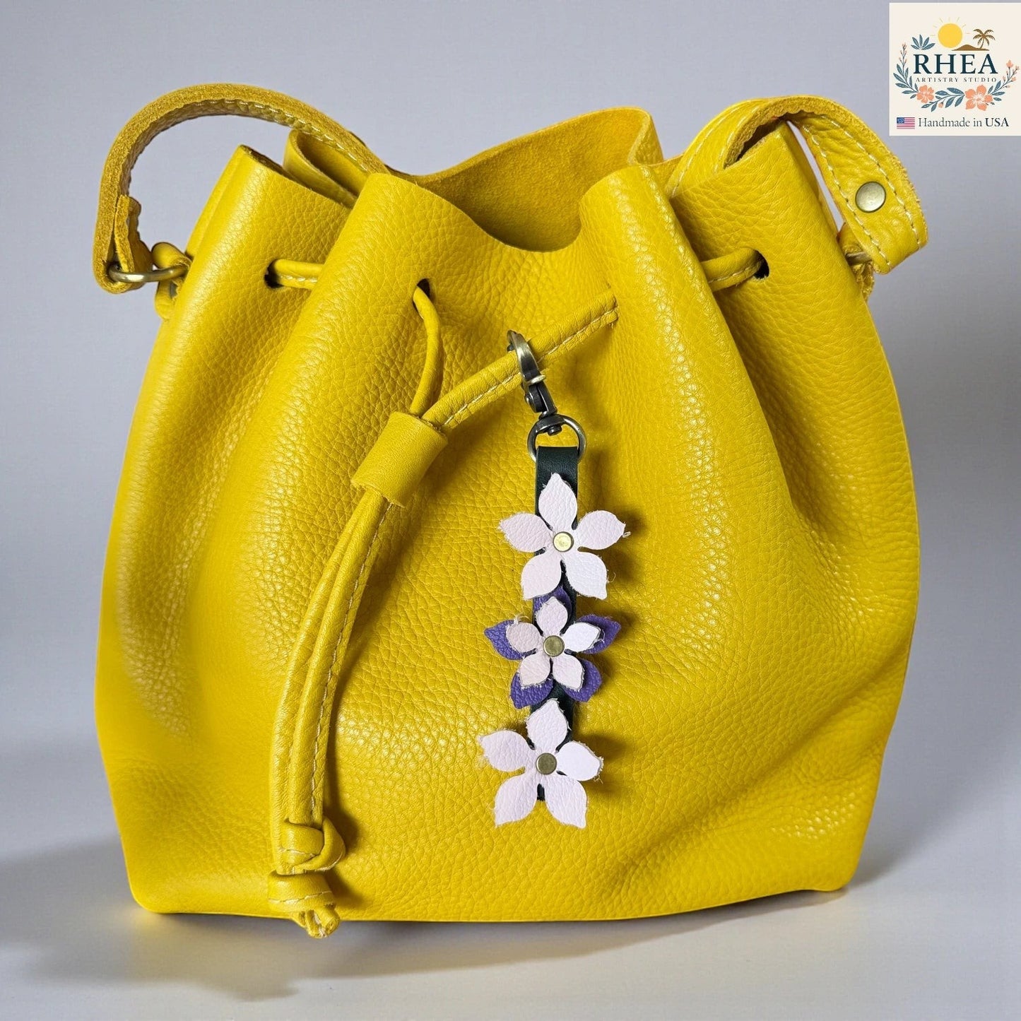 Rhea Artistry Studio yellow leather bucket bag with a handmade leather floral charm featuring light pink and purple flowers and a brushed brass clasp.