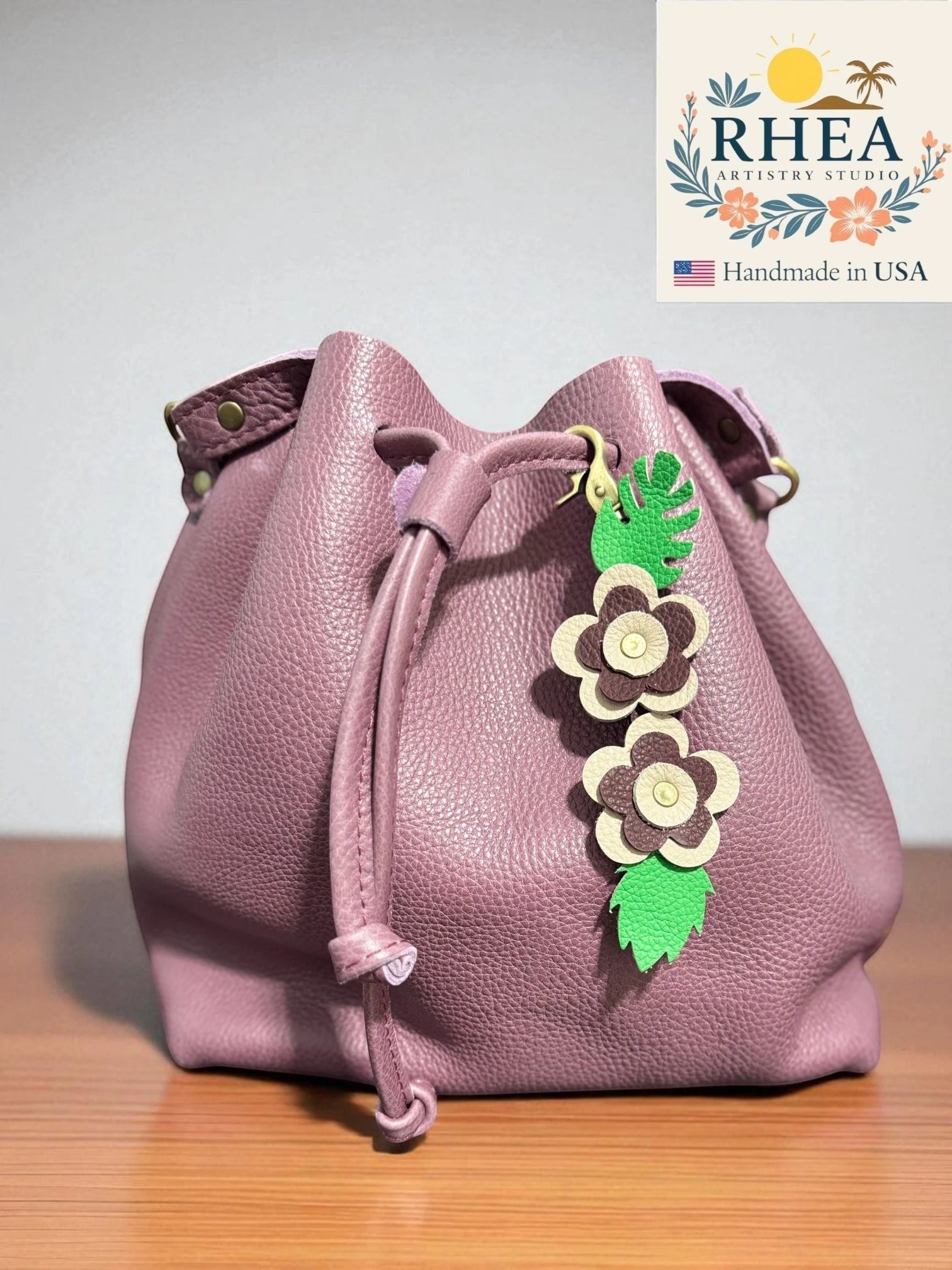 Rhea Artistry Studio handmade leather floral keychain charm with green leaves and beige and brown flowers, attached to a dusty rose bucket bag.