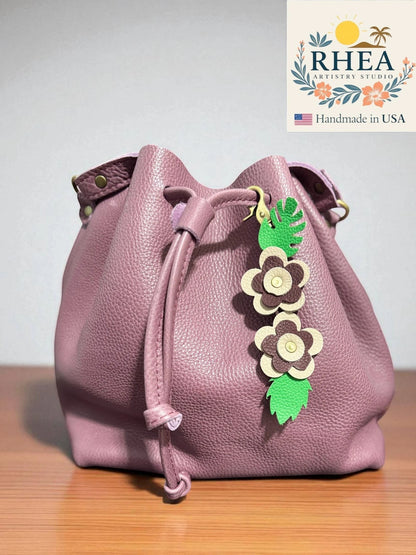 Rhea Artistry Studio handmade leather floral keychain charm with green leaves and beige and brown flowers, attached to a dusty rose bucket bag.
