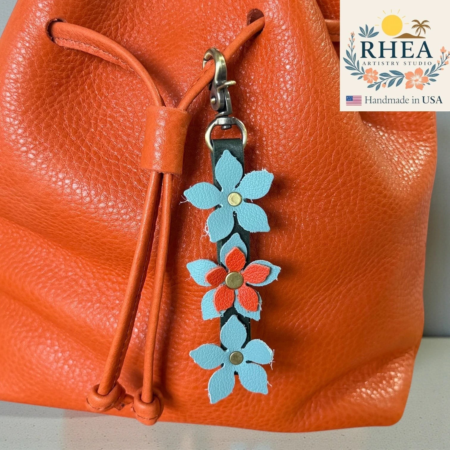 Rhea Artistry Studio handmade leather floral charm with light blue and coral flowers and brushed brass clasp attached to an orange leather bag.