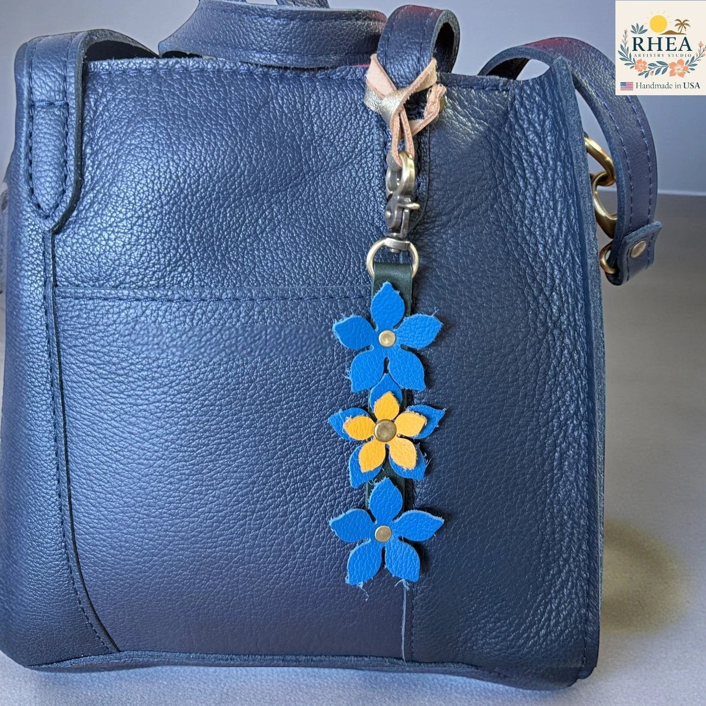 Rhea Artistry Studio handmade leather floral charm with blue and yellow flowers and a brushed brass clasp attached to a navy blue handbag.