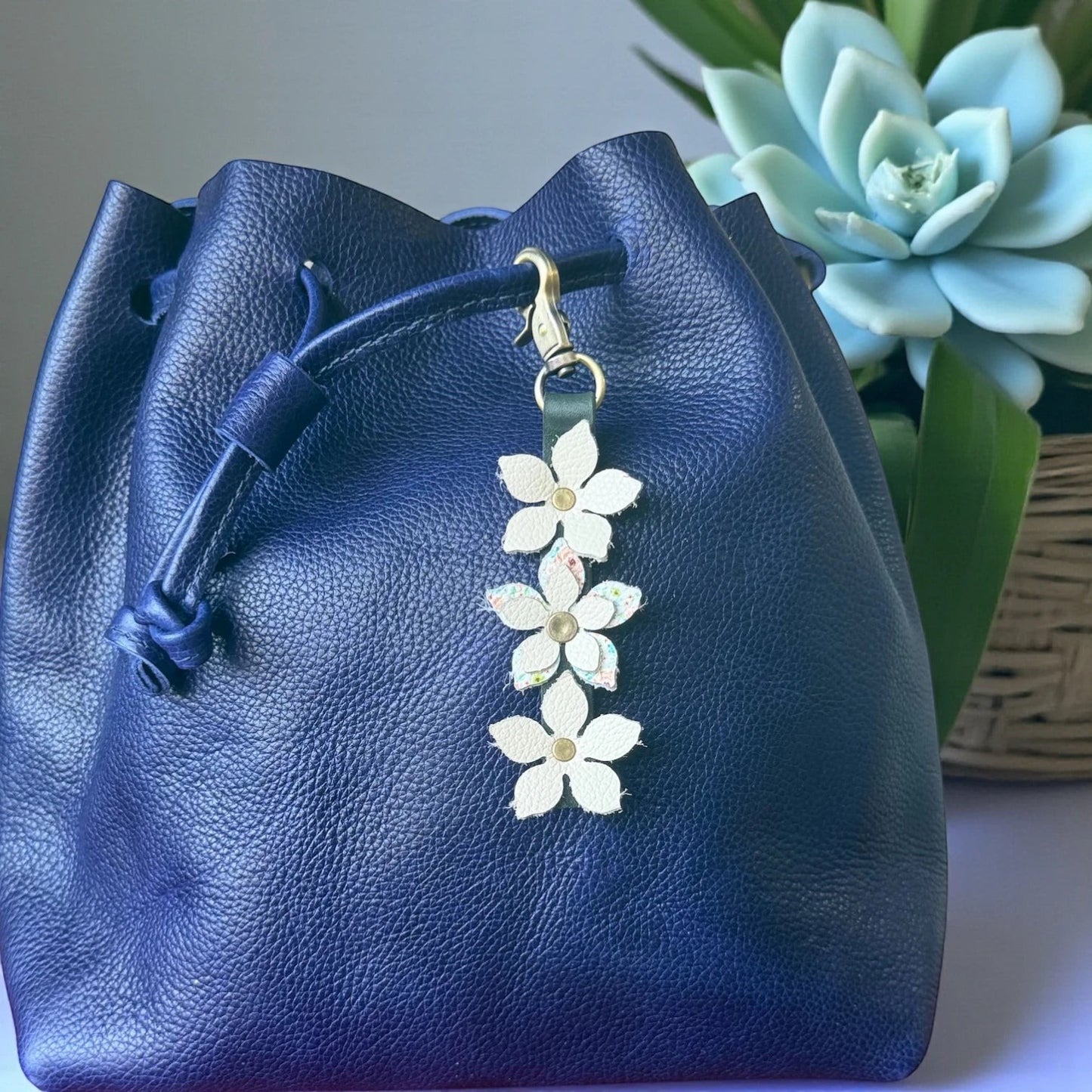 Rhea Artistry Studio navy blue leather drawstring bag with a handmade floral charm featuring white leather flowers and a brushed brass clasp.