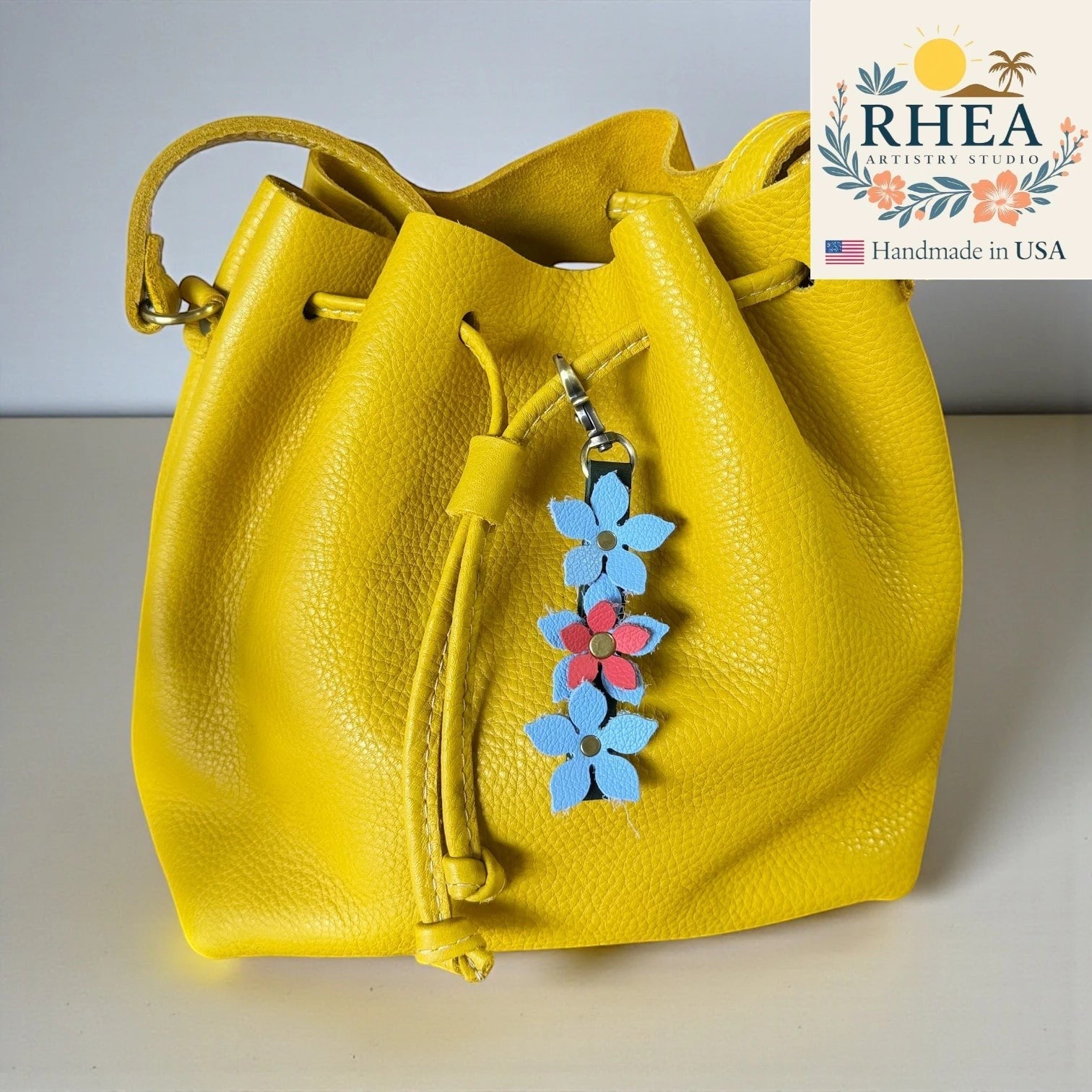 Rhea Artistry Studio handmade leather floral charm with blue and pink flowers and a brushed brass clasp, attached to a yellow leather bucket bag.