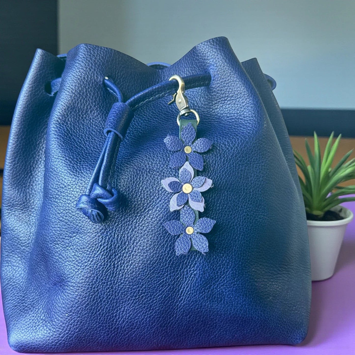 Rhea Artistry Studio navy blue leather bucket bag with a handmade floral charm featuring purple leather flowers and a brushed brass clasp.