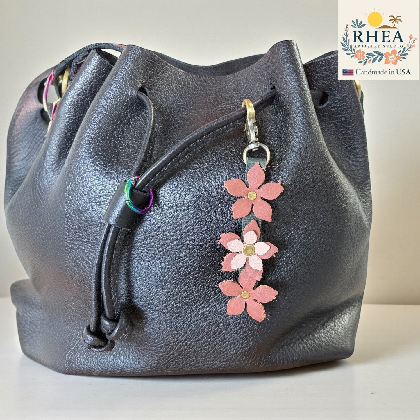 Rhea Artistry Studio handmade leather floral charm with brushed brass clasp, featuring three pink and light pink flowers on a dark grey leather bucket bag.