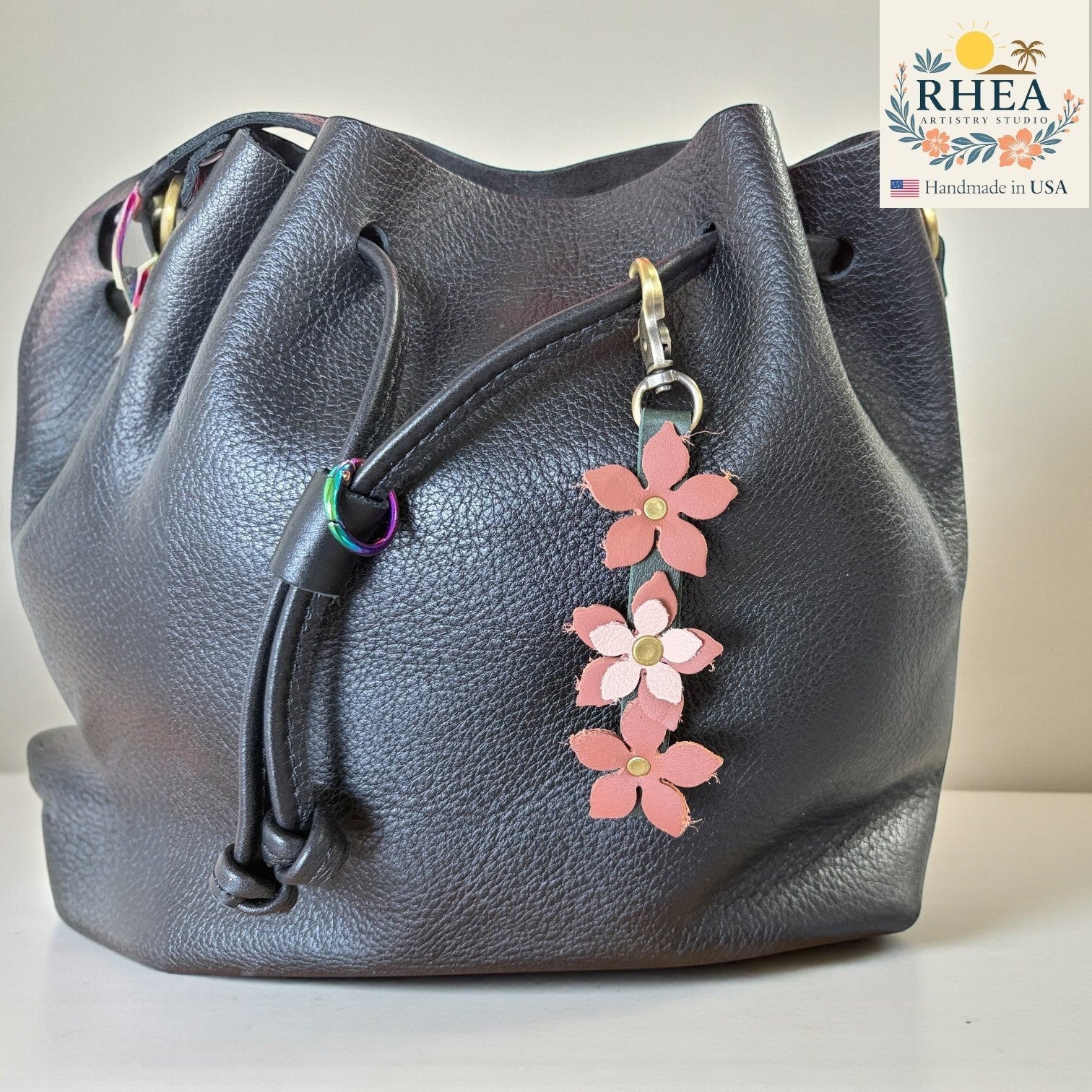 Rhea Artistry Studio handmade leather floral charm with brushed brass clasp, featuring three pink and light pink flowers on a dark grey leather bucket bag.