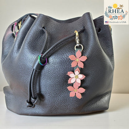 Rhea Artistry Studio handmade leather floral charm with brushed brass clasp, featuring three pink and light pink flowers on a dark grey leather bucket bag.