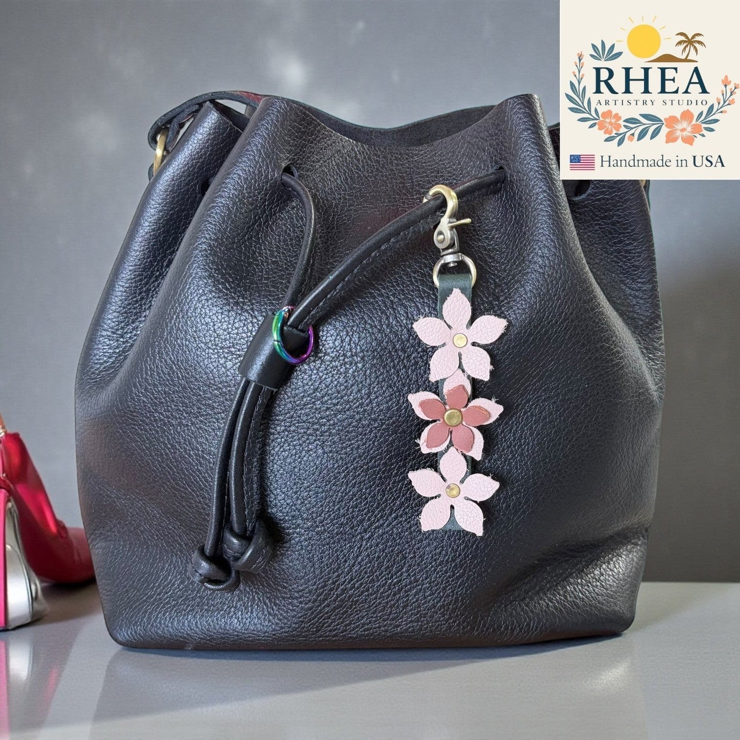 Rhea Artistry Studio handmade leather floral charm with pink and mauve flowers and a brushed brass clasp, attached to a black leather bucket bag.