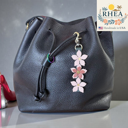 Rhea Artistry Studio handmade leather floral charm with pink and mauve flowers and a brushed brass clasp, attached to a black leather bucket bag.